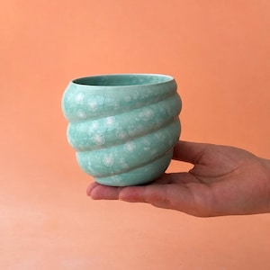 Puede incluir: Un pequeño recipiente de cerámica turquesa con un diseño en espiral se sostiene en una mano. El recipiente tiene una superficie texturizada con patrones florales blancos. El fondo es de color melocotón. El recipiente mide unos 10 cm de ancho y 8 cm de alto.