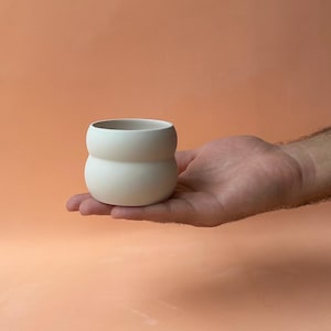 Könnte beinhalten: Ein kleines, cremefarbenes Keramikgefäß mit einer abgerundeten, sanduhrartigen Form. Die glatte, matte Oberfläche kontrastiert mit dem pfirsichfarbenen Hintergrund. Das Gefäß wird in der offenen Hand einer Person gehalten.