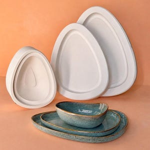 Könnte beinhalten: Eine Sammlung weißer, tropfenförmiger Servierplatten und Schalen. Die weißen Platten haben einen erhöhten Rand. Eine türkisfarbene Schüssel und zwei passende Platten mit Goldrand sind ebenfalls enthalten. Die Artikel sind auf einer pfirsichfarbenen Oberfläche angeordnet.