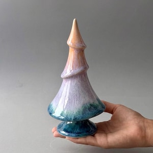 May include: A ceramic tree-shaped sculpture with a glossy finish, featuring a color gradient that transitions from a deep blue base to a soft lavender middle section and ends with a pale peach-colored pointed top, standing on a circular base.