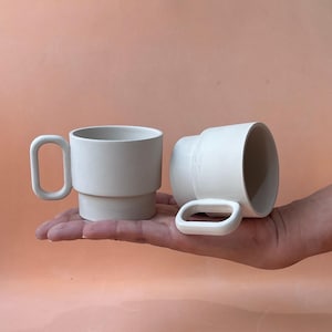 Peut inclure: Deux tasses en céramique blanche au design minimaliste. L'une est droite, l'autre est couchée. Chaque tasse a une forme de poignée unique. Les tasses sont présentées sur un fond pêche.