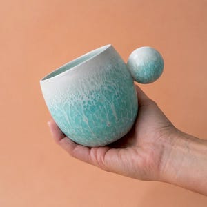 以下が含まれることがあります： ユニークな球状のハンドルが付いたセラミックマグ。マグカップは、縁の白からターコイズの色合いに変わり、テクスチャ加工された大理石模様の効果があります。ハンドルは、お揃いのターコイズ色の球体です。マグカップは、桃色の背景に対して保持されています。