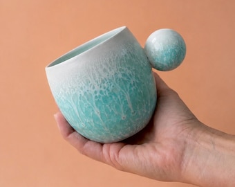 May include: A ceramic mug with a unique spherical handle. The mug transitions from white at the rim to a turquoise hue, with a textured, marbled effect. The handle is a matching turquoise sphere. The mug is held against a peach-colored background.