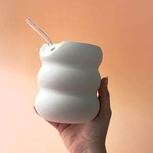以下が含まれることがあります： 白く有機的な形のセラミックカップと透明なガラスストロー。カップはユニークで丸みを帯びたデザインで、桃色の背景に配置されています。ストローはカップの側面の穴に差し込まれています。