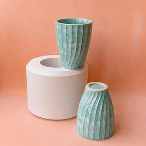 May include: Two turquoise ceramic cups with a textured, ribbed design. One cup is upright, the other is inverted. They are displayed on a white cylindrical stand against a peach background. The cups have a speckled pattern.