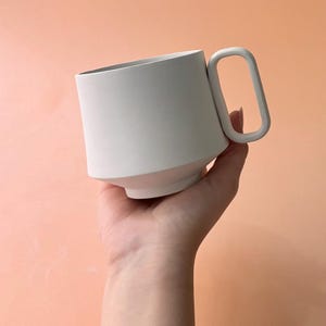 Könnte beinhalten: Eine weiße Keramik-Tasse mit einem einzigartigen, kantigen Design. Die Tasse hat eine geometrische Form und einen rechteckigen Griff. Sie wird vor einem pfirsichfarbenen Hintergrund gehalten, der ihre minimalistische Ästhetik hervorhebt. Die Tasse ist eine stilvolle Ergänzung für jede Küche, mit einem Fassungsvermögen von ca. 300 ml.