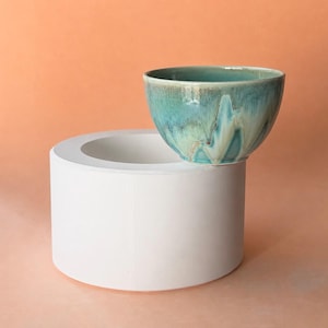 May include: A turquoise ceramic bowl with a dripped glaze effect sits atop a white cylindrical pedestal. The bowl's interior and exterior feature shades of blue, green, and brown. The background is a solid peach color.