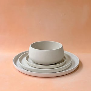 May include: A set of four light gray ceramic dishes. The set includes a large, shallow plate, two smaller plates, and a rounded bowl. The dishes are stacked on top of each other, with the bowl at the top. The background is a solid peach color.