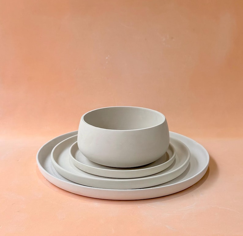 May include: A set of four light gray ceramic dishes. The set includes a large, shallow plate, two smaller plates, and a rounded bowl. The dishes are stacked on top of each other, with the bowl at the top. The background is a solid peach color.