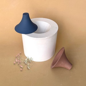 May include: Two silicone funnels, one blue and one brown, are sitting on a white cylindrical base. The base has a circular opening at the top. A small bunch of dried flowers is in the foreground.