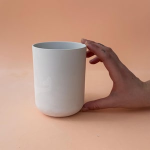 Puede incluir: Un jarrón cilíndrico de cerámica blanca con base redondeada. La mano sostiene el jarrón sobre un fondo color melocotón. El jarrón tiene un acabado liso y mate y un diseño sencillo y moderno.