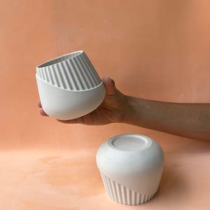 May include: Two white ceramic vases with a contemporary design. One vase is held, tilted, and the other is upright. Both have a smooth, rounded base and a section with vertical ridges. The background is peach.