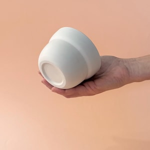 Puede incluir: Una maceta de cerámica blanca con forma redondeada, sostenida en una mano. La maceta tiene una base ancha y una parte superior ligeramente ensanchada, con un acabado liso y mate. El fondo es de color melocotón sólido.