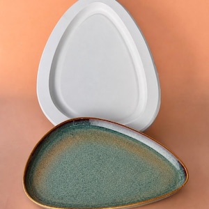 May include: Two decorative serving trays. The top tray is white and has a raised, rounded triangular shape. The bottom tray is teal with a brown rim. The trays are on a peach-colored background. The trays are likely for serving food or displaying items.