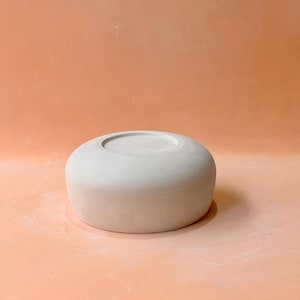 May include: A light gray, ceramic bowl with a rounded shape and a recessed center. The bowl is smooth and sits on a peach-colored surface. The design is simple and modern, suitable for various uses.