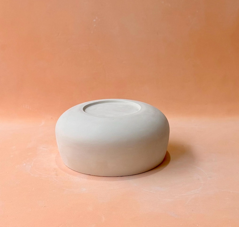 May include: A light gray, ceramic bowl with a rounded shape and a recessed center. The bowl is smooth and sits on a peach-colored surface. The design is simple and modern, suitable for various uses.