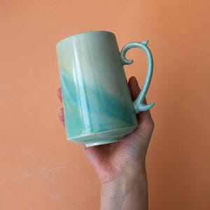 Könnte beinhalten: Eine Keramik-Tasse mit einem einzigartigen Henkel-Design. Die Tasse hat einen Farbverlauf von hellgrün zu Türkis. Der Henkel ist geschwungen und hat ein kleines dekoratives Element am Ende. Die Tasse ist etwa 10 cm hoch.