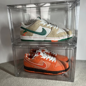 May include: Two pairs of Nike trainers in a clear plastic shoe box. The top pair is a light brown and green colour with a white swoosh. The bottom pair is orange and black with a white swoosh.