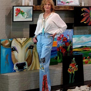May include: An array of framed paintings, featuring floral and animal subjects, are displayed on a brick wall. The paintings showcase a variety of styles and colors. The woman is wearing light blue jeans with patches and a white blouse.