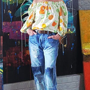 May include: An off-the-shoulder blouse with a citrus fruit pattern, paired with distressed blue jeans and brown clogs. The blouse features orange and green fruit designs on a white background. The jeans have patches and a brown belt.