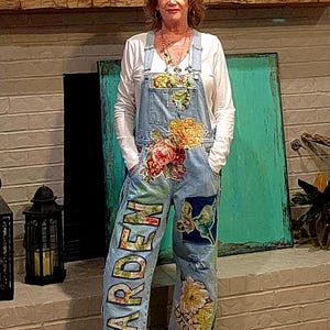 May include: Light blue denim overalls with floral and butterfly designs. The word "GARDEN" is painted on the left leg. The right leg has the word "Dream" and a floral design. The overalls are worn over a white long-sleeve shirt.