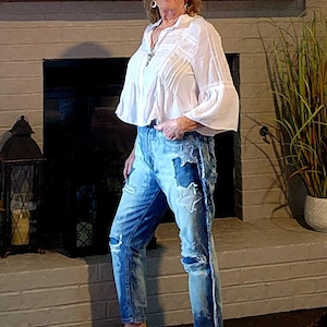 May include: A woman wearing a white blouse with bell sleeves and distressed blue jeans with star patches. She is wearing brown slip-on shoes. The background includes a fireplace and decorative items.