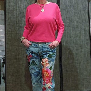 May include: A woman wearing a long-sleeved, bright pink top and light blue jeans. The jeans feature a floral design in shades of red, pink, and orange. She is also wearing a gold necklace and bracelet.