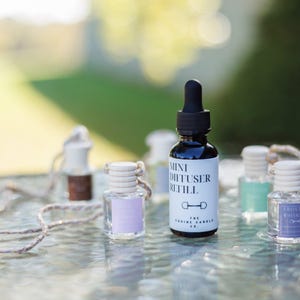 May include: A collection of mini diffuser refill bottles in various colors, including a black bottle with a dropper and a white label. The bottles are arranged on a reflective surface with a blurred green background. The text "Mini Diffuser Refill" is visible on the label.