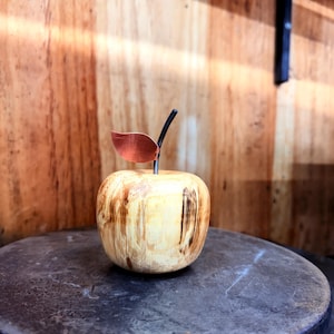 Könnte beinhalten: Eine Holzapfel-Skulptur mit einem Kupferblatt und einem schwarzen Stiel. Der Apfel ist hellbraun mit dunkleren braunen Streifen und steht auf einer dunklen Metalloberfläche. Der Hintergrund ist eine Holzwand.