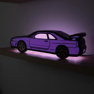 May include: A purple car-shaped lamp with a black outline and gold wheels sits on a wooden shelf. The lamp emits a soft purple glow, illuminating the car's interior and the shelf below. The lamp is plugged into an electrical outlet.