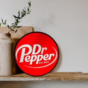May include: A round Dr Pepper sign with a red background and white lettering. The sign features the Dr Pepper logo and the text "Est. 1885". The sign is displayed on a wooden shelf.