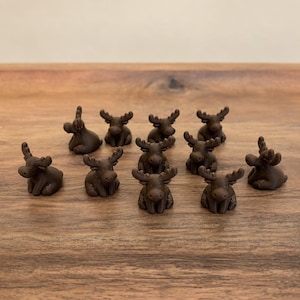 May include: A collection of ten small, dark brown moose figurines arranged in a group. Each miniature moose features a rounded body, a distinct head with prominent antlers, and a seated posture, all crafted with a uniform matte finish.