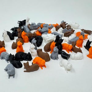 May include: A collection of small, 3D printed cat figurines in various colors, including black, white, gray, orange, and brown. The figurines are all in different poses, some sitting, some standing, and some lying down.