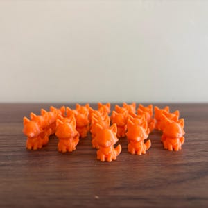 May include: A collection of small, orange fox figurines arranged on a dark wooden surface. The figurines are uniformly colored and detailed, with visible ears, tails, and facial features. The background is a neutral, light-colored wall.
