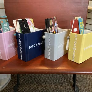May include: Four colorful bookends shaped like books with the word "BOOKMARKS" printed on the spine. Each bookend has a bookmark attached to it. The bookmarks are different colors and have different designs.