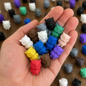 May include: A collection of small, three-dimensional owl figurines in various solid colors including red, yellow, blue, green, purple, white, brown, black, and gray. Each owl features pointed ears, large circular eyes, and a small beak.