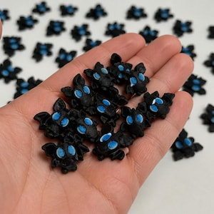 May include: A collection of small, black, bat-shaped charms featuring prominent, bright blue circular eyes. These miniature figures have textured surfaces and are shaped like stylized bats with pointed ears and small wings, presented in a group.