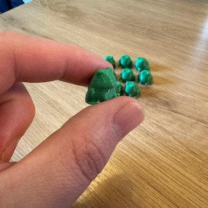 May include: A group of small green frog figurines, one of which is being held by a hand.