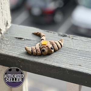Könnte beinhalten: Handgefertigte Ton-Skulptur eines Croissants mit Butter. Die Skulptur ist hellbraun mit dunkelbraunen Details und einem kleinen gelben Butterstück. Die Augen sind schwarz. Auf einer verwitterten Holzoberfläche platziert.
