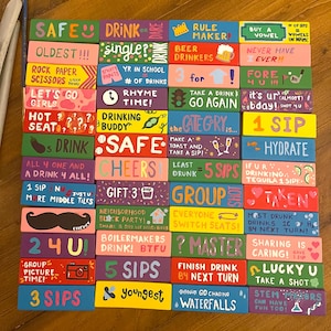 May include: A set of colorful wooden blocks with various drinking game rules and challenges written on them. The blocks are painted in bright colors with black outlines and feature illustrations such as a beer mug, a golf club, a house, and a mustache.