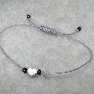 Puede incluir: Una delicada pulsera de cordón lavanda con un dije de corazón plateado y pequeñas cuentas burdeos. La pulsera es ajustable con un cierre de nudo corredizo. Una pieza de joyería sencilla y elegante.