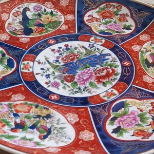 May include: A large, round, white ceramic plate with a blue and red border. The plate is decorated with a floral pattern and peacocks. The plate is a traditional Japanese design.