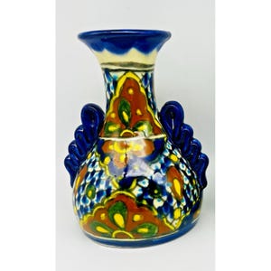 May include: A ceramic vase featuring a flared rim and two decorative side handles. The surface is decorated with a vibrant hand-painted pattern in shades of deep blue, burnt orange, yellow, and green, incorporating floral motifs and grid designs.
