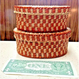 May include: Two round, lidded woven baskets stacked vertically, featuring a repeating pattern of alternating rust-orange and natural beige fibers. A United States one-dollar bill is placed in front of the baskets to provide a scale reference.
