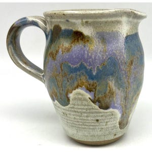 May include: A ceramic pitcher featuring a rounded body and a curved handle. The surface displays a textured, off-white base with a flowing glaze pattern in shades of lavender, blue, and brown that drips down from the rim toward the base.