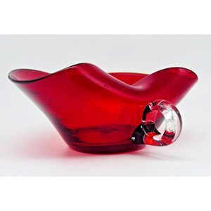 Könnte beinhalten: Eine leuchtend rote Glasschale mit einem breiten, gewellten Rand und einer glatten, durchscheinenden Oberfläche. Die Schale verfügt über einen einzelnen, klaren Glasgriff an einer Seite, der eine abgerundete, massive Form aufweist und einen Kontrast zum tiefroten Körper bildet.