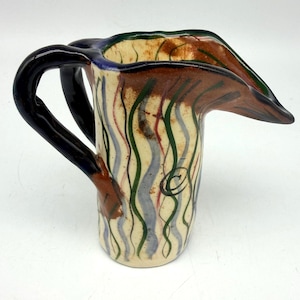 May include: A ceramic pitcher with a cream-colored base featuring vertical wavy lines in green, blue, and brown. The pitcher has a dark blue handle, a wide pouring spout, and a hand-painted appearance with a visible copyright symbol on the side.