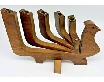 Puede incluir: Un candelabro de madera con forma de pájaro y cinco brazos verticales, cada uno rematado con una pequeña copa de latón para velas. La madera marrón muestra una veta visible y la estructura se apoya en una base central inferior.