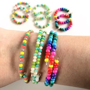 May include: A set of colorful beaded bracelets, each with a different color scheme. The bracelets are made with small, round beads and are strung on a silver wire. The bracelets are stacked on a person's wrist.