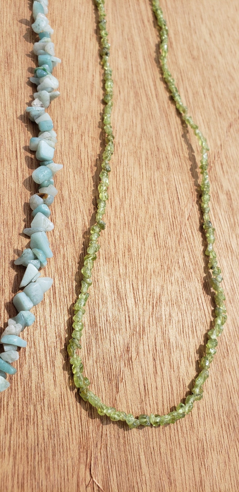 May include: Two necklaces made of natural stone beads. One necklace is made of light blue aquamarine beads and the other is made of green peridot beads. The necklaces are displayed on a wooden surface.